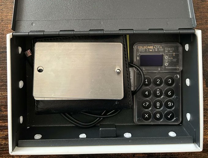 A Coldcard Mk4 hardware wallet (branded "COLDCARD") and its accompanying metal seed phrase backup plate are shown neatly stored inside a cardboard box. The image illustrates the article's point about cold wallet security vulnerabilities, depicting the very type of device involved in the reported $130 million hack stemming from a 2021 update bug.