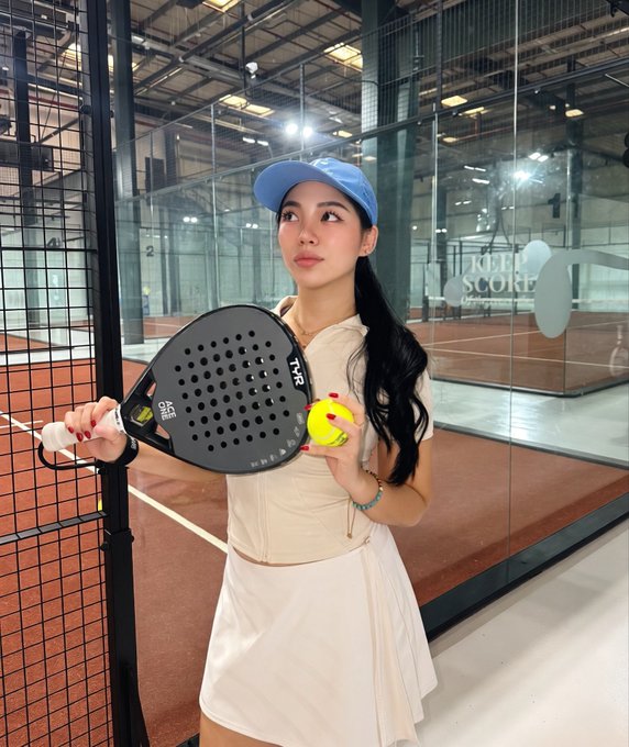 A young woman in athletic wear and a blue visor holds a padel racket and ball in an indoor sports facility.