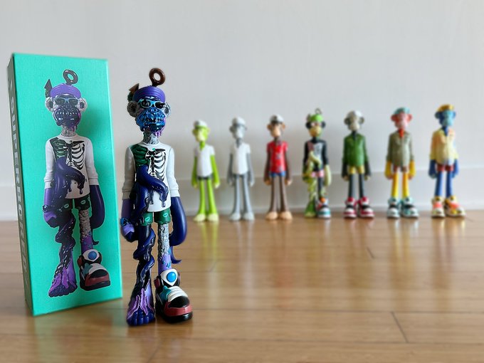 A row of colorful collectible skeleton figurines stands on a wooden floor next to its mint-green packaging box featuring a skeleton character with sunglasses.