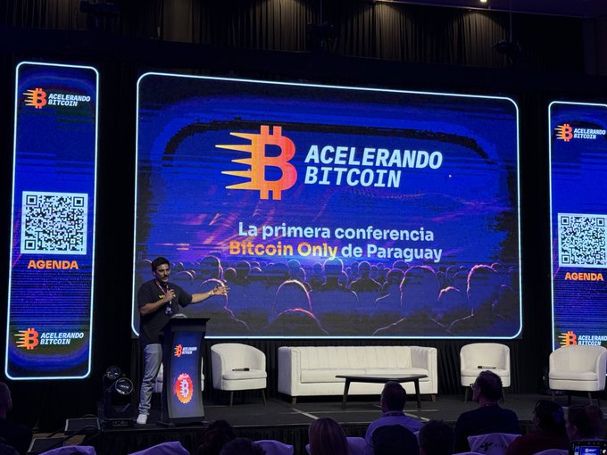 A man speaking at a podium on a stage in front of a large screen displaying 'ACELERANDO BITCOIN' and 'La primera conferencia Bitcoin Only de Paraguay', with an audience visible in the foreground.
