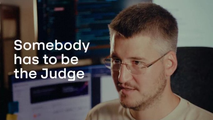 A man wearing glasses smiles slightly in what appears to be a workspace, with the text "Somebody has to be the Judge" overlaid on the left side of the image.