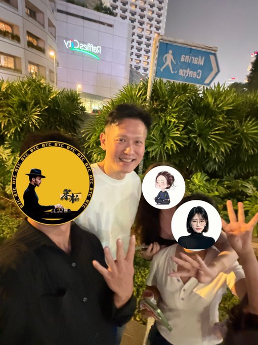 A group photo taken at night in front of a Desigual store, featuring a man smiling in the center and other people with their faces covered by BTC coin graphics and avatar stickers, posing with peace signs.