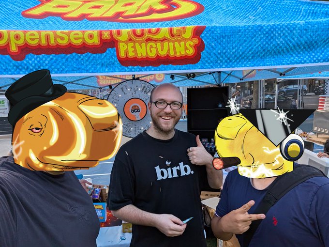 A photo of three people standing under a 'Pudgy Penguins' branded tent, with the person in the middle wearing a 'birb.' shirt and giving a thumbs up; the faces of two people are covered with cartoon emojis.