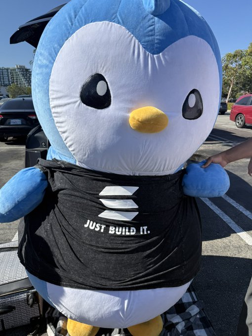 A giant inflatable Pengu mascot stands outdoors wearing a black shirt that reads "JUST BUILD IT." with a Solana logo, promoting the Solana Summer House event in LA on August 29th.