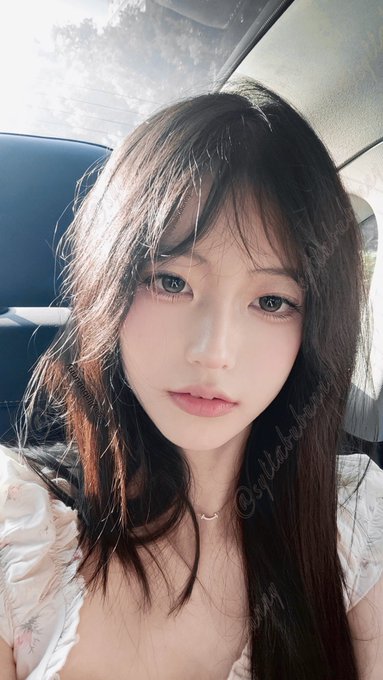 A close-up selfie of a young woman with long dark hair sitting in a car, with a watermark '@sylviakoo_' visible on the image.