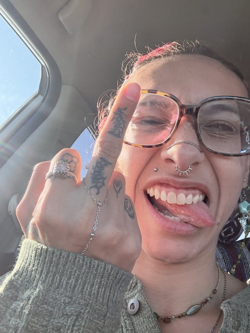 A person with glasses, a nose septum piercing, and visible hand tattoos smiles playfully while sticking their tongue out and holding up a middle finger in a car.