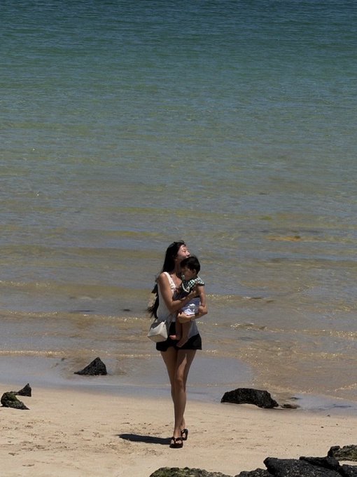 A photograph of a woman holding a child while walking along a sandy beach near the water.