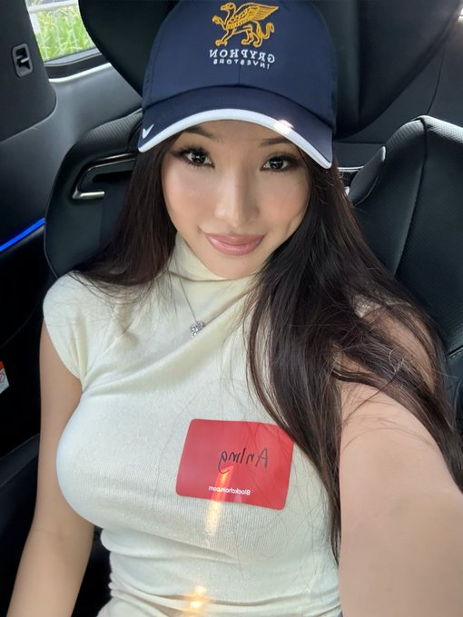 A woman wearing a navy blue 'Gryffindor Quidditch' baseball cap and a cream-colored sleeveless top takes a selfie inside a car, with a red 'Milano BlackBox Inc.' sticker visible on her clothing.