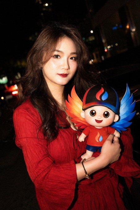 A young woman with long dark hair wearing a red dress holds a winged mascot doll with a flame logo on its hat and shirt, photographed at night in a city setting.