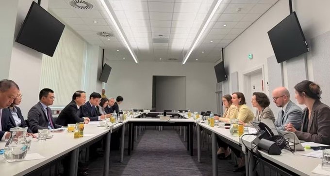 A formal meeting takes place in a conference room with two long tables where men in dark suits on the left face a group including women and men on the right, with water glasses, microphones, and wall-mounted screens visible.