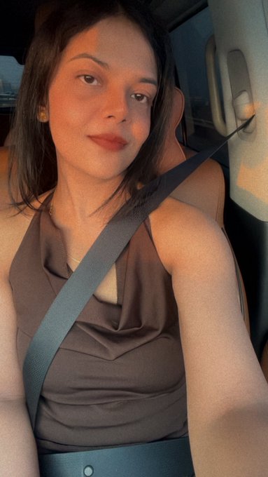A young woman with long dark hair sits in the passenger seat of a car, wearing a brown halter top with a seatbelt strapped across her chest, illuminated by warm sunset light. The cozy, golden-hour selfie complements the post's friendly greeting, suggesting she's in Dubai ready to connect with fellow crypto and forex enthusiasts.