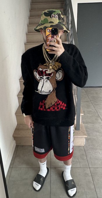A person takes a mirror selfie on a staircase wearing a camo bucket hat, gold chains, a black sweater featuring a cartoon monkey graphic, and black basketball shorts with slide sandals.