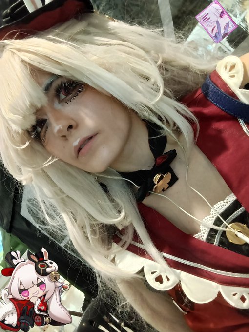 A close-up selfie of a person in cosplay wearing a white wig, dramatic makeup, and a red and white outfit with a black choker, with a small cartoon avatar sticker in the corner.