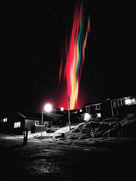 A nighttime photograph of a snowy village under a starry sky featuring a striking, multicolored aurora-like light display in red, green, and blue streaming down toward the houses.