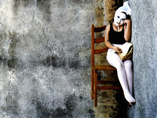 A surreal photo of a person wearing a white bunny mask and bodysuit sitting on a wooden chair attached to a weathered concrete wall.