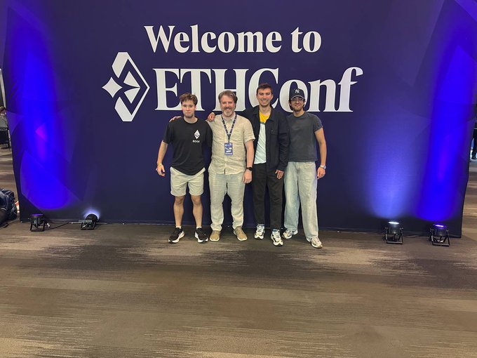 The image features four young men posing for a photo in front of a blue wall that displays "Welcome to EthConf" written in white letters. The group appears to be attending an event or gathering related to Ethereum technology, as indicated by the presence of a sign on the wall. They are standing close together and smiling, creating a sense of camaraderie among them.