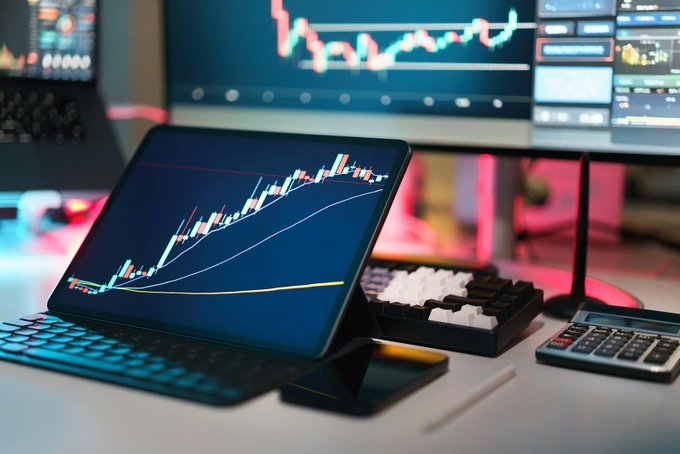 Trading setup with a tablet and monitor displaying candlestick charts with red and green candles, accompanied by a mechanical keyboard and calculator on a desk.