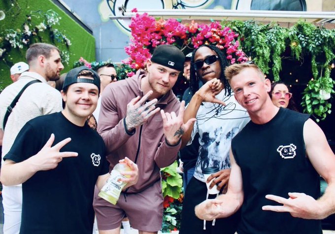 A group photo of four men posing together at what appears to be a social event with flowers in the background, with two wearing matching black shirts featuring a logo.