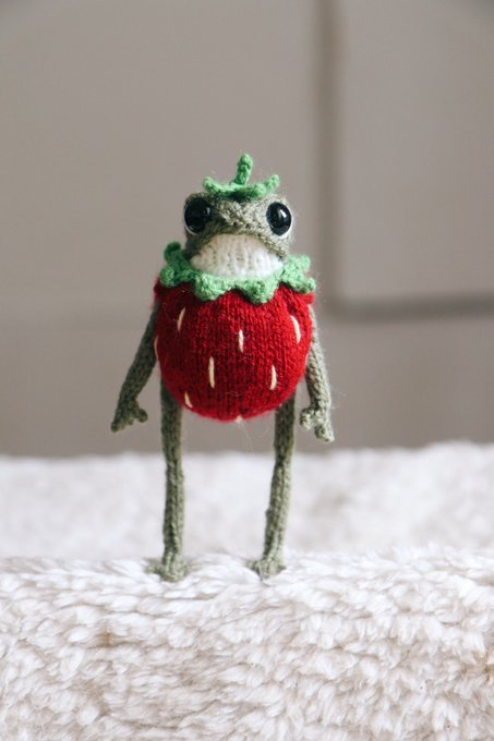 A small crocheted amigurumi figure of a green frog sitting inside or atop a red strawberry, with yarn arms and legs dangling down on a white surface.