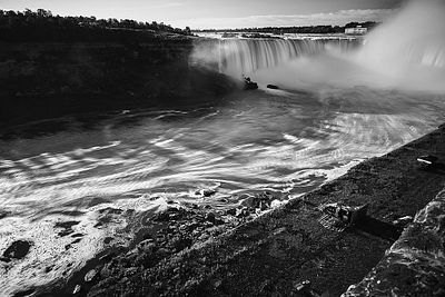A black and white photograph of a powerful waterfall cascading into a river, with mist rising from the water and a small boat visible near the falls.