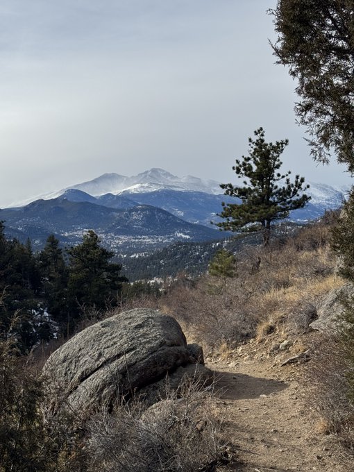 Scenic landscape photograph taken from a hiking trail, showing a rocky dirt path in the foreground with a view of snow-capped mountains and pine trees in the distance.