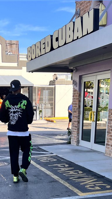 A person wearing a black hoodie with green graphics, black pants with green designs, and green shoes walking toward a storefront with a "BORED CUBAN" sign.