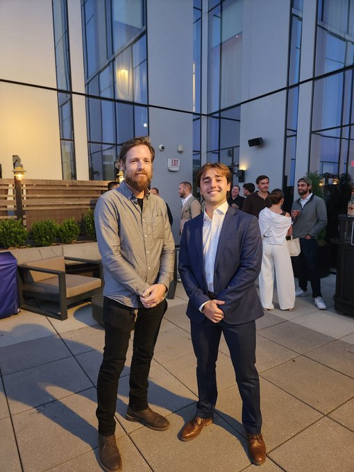 Two men pose for a photo at an outdoor evening gathering; the man on the right is wearing a navy suit, while the man on the left has a beard and is wearing a blue shirt.