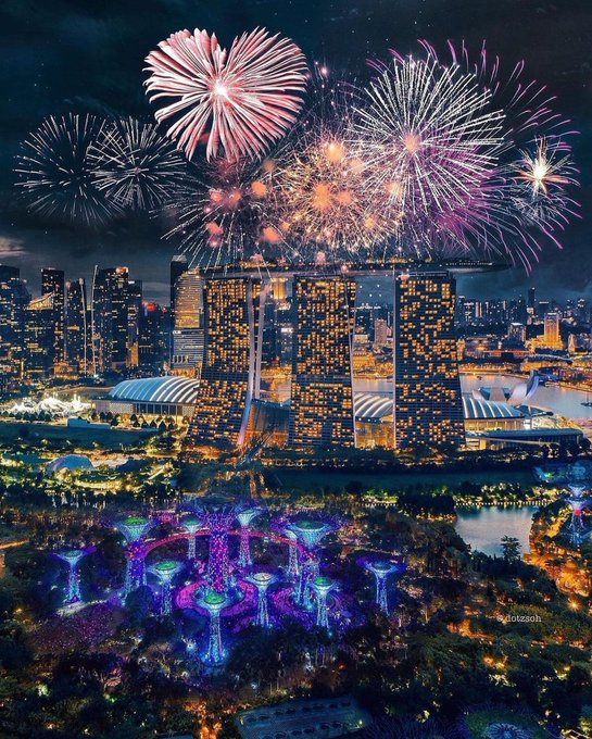 A stunning aerial night view of Singapore's Marina Bay Sands lit up with golden windows, surrounded by vibrant Supertree Grove displays and a spectacular fireworks show overhead — capturing the celebratory spirit of July 4th, coinciding with America's 250th birthday. Watermarks "dotsash" are visible in the lower right.
