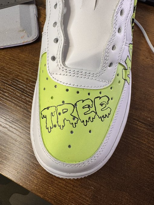 A white high-top sneaker with a neon green toe box featuring the word 'TREE' in a dripping, graffiti-style black font rests on a wooden surface.