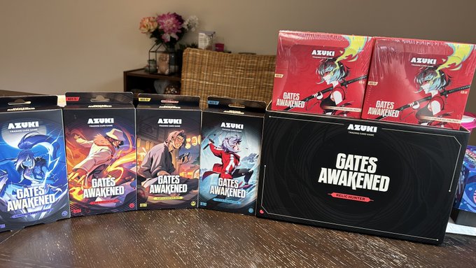 A collection of "Azuki Elements Collection" and "Gates Awakened" TCG card packs, blister packs, and a storage tin are displayed on a table in celebration of the Azuki TCG launch day.