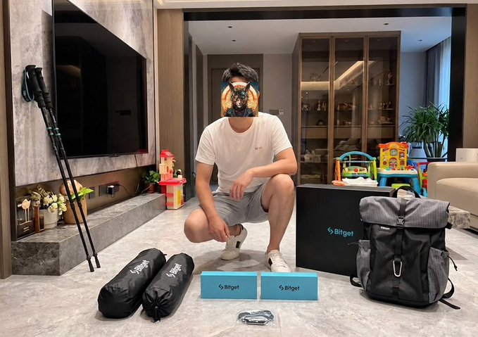 A man wearing a dog mask squats in a modern living room displaying a Bitget product lineup, including a branded box, backpack, sleeping bag, and camping chairs.