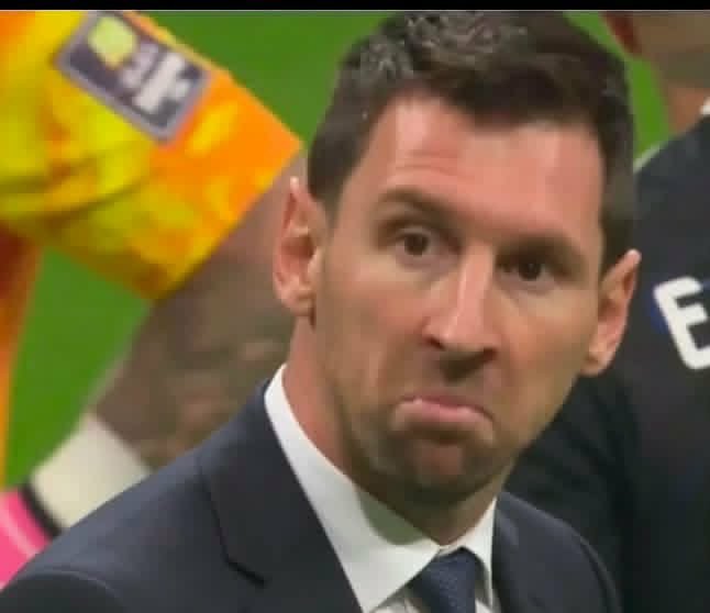A close-up photo of soccer player Lionel Messi wearing a dark suit and tie, making a slightly pouting facial expression with a colorful background behind him.
