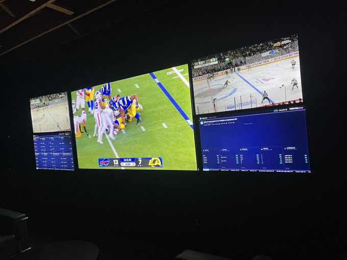 Multiple screens displaying live sports broadcasts, with the center monitor showing a football game between Buffalo (13) and the Los Angeles Rams (7) in the first quarter, flanked by what appears to be hockey games on adjacent displays.