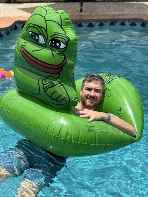 The image features a man wearing a green inflatable frog costume while swimming in a pool, with his head above water. The caption of the image reads "I sold meme coins & built a pool Shout out to @Based_AllMight for the $PEPE tube!".