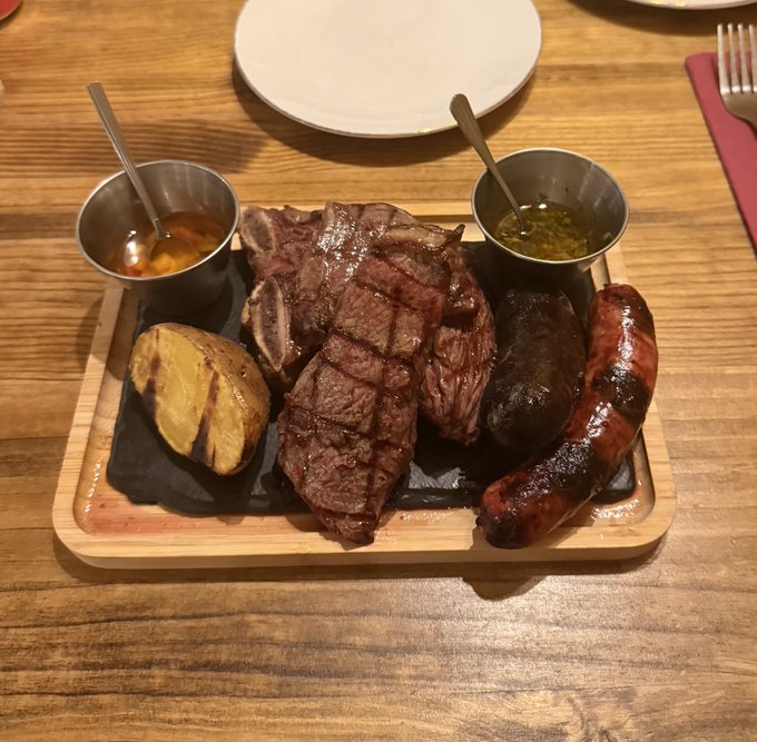 A wooden serving board featuring various grilled meats including a steak and sausage, accompanied by a baked potato and two small metal cups containing sauces.
