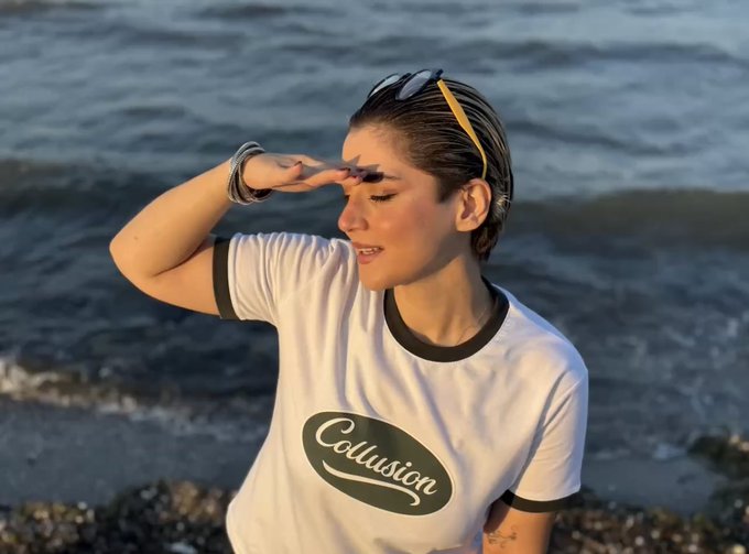 A young woman with wet hair, sunglasses on her head, stands by the ocean wearing a white 'Collusion' ringer t-shirt, shielding them from the light.