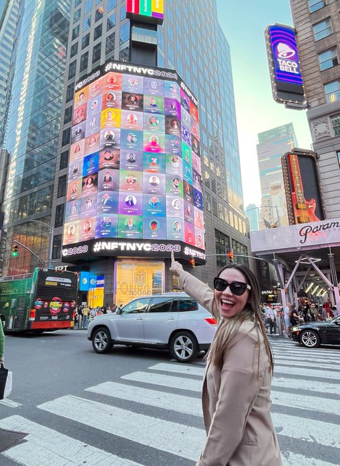 A woman points excitedly at a Times Square billboard in New York City displaying "#NFTNYC2023" alongside dozens of profile portraits. Her joyful expression matches her caption about being genuinely surprised to find her face featured on the iconic digital display during the NFT NYC event.