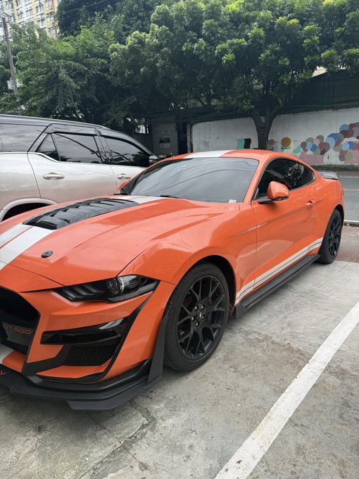 An orange Ford Mustang GT500 with black stripes and racing accents parked in a lot next to other vehicles, with trees in the background.