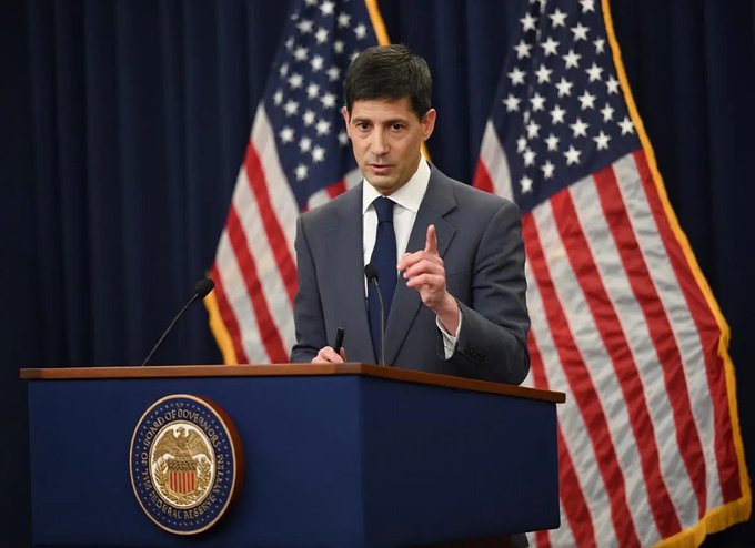 A man in a suit stands at a podium bearing the U.S. Board of Governors seal, speaking in front of two American flags. The image pairs with the post about favorable U.S. CPI inflation data, visually evoking a Federal Reserve official responding to the bullish economic news.
