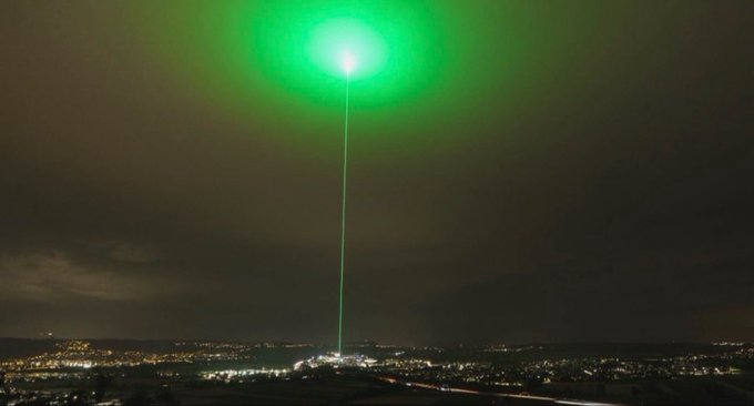 A vibrant green laser beam shoots straight up into the night sky, illuminating the clouds in a striking emerald glow above a cityscape. The image serves as a dramatic visual metaphor for a memecoin about to "moon"—symbolizing an explosive upward price trajectory, much like the laser piercing through the darkness to reach new heights.