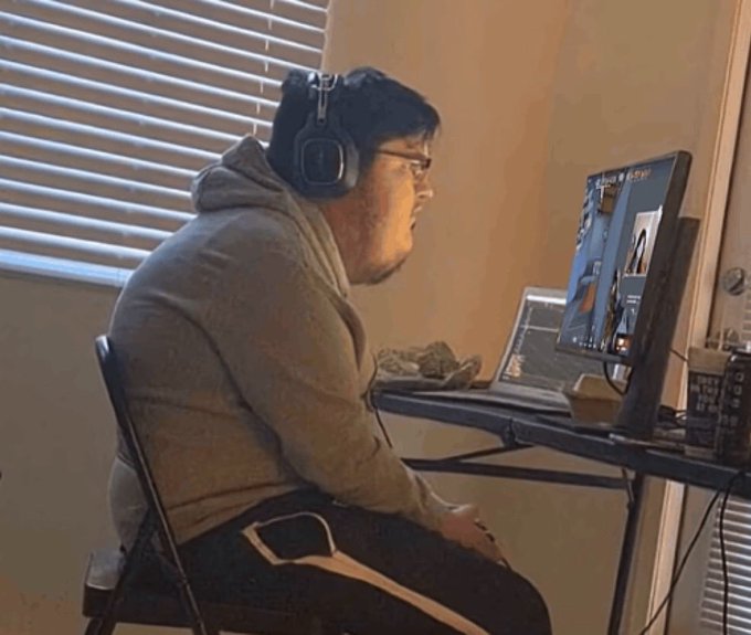 A man wearing a gray hoodie and large black headphones sits in front of a laptop and monitor, appearing focused on the screen which displays what looks like a video or image editing interface.