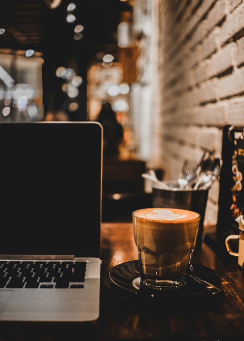 A dimly lit café scene featuring an open laptop next to a latte with heart-shaped latte art on a wooden table, set against a rustic exposed brick wall.