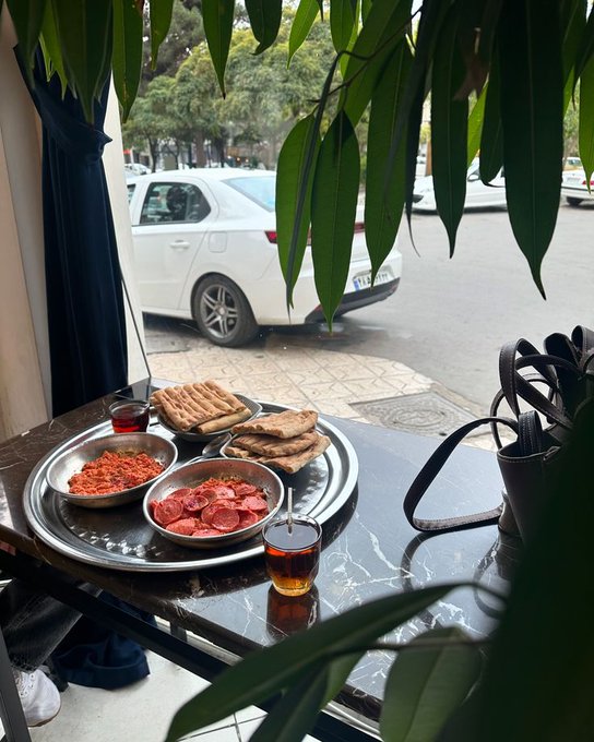 A breakfast spread of flatbread, sliced tomatoes, and a red dip served on metal plates at a cafe table, with a white car parked on the street visible through the window.