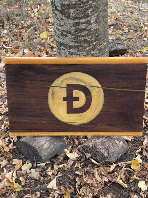 A wooden box or sign with a circular gold inlay featuring a stylized "D" logo, placed outdoors against a tree trunk with fallen leaves.