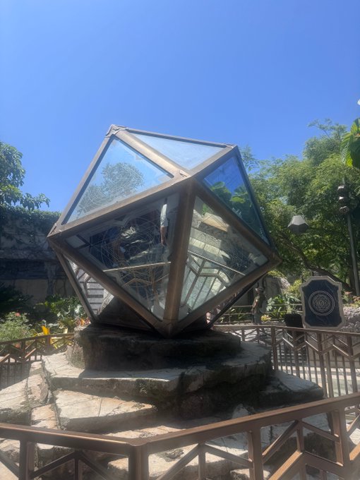 A large geometric polyhedron sculpture made of glass panels with a dark metal frame, displayed outdoors on a stone pedestal surrounded by a metal fence, with lush green trees and clear blue sky in the background.