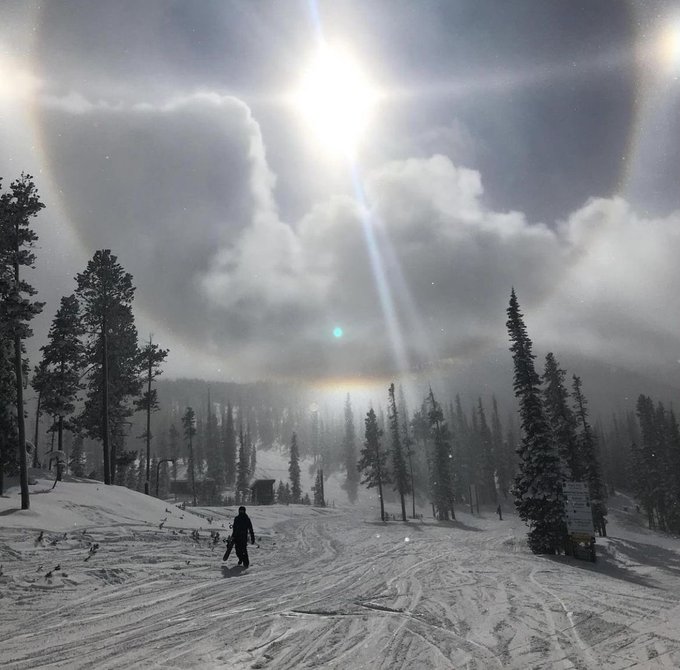 A snowboarder rides down a snowy mountain slope under a bright sun surrounded by dramatic clouds, with evergreen trees lining the trail and a sign visible on the right. The image reflects the "let's make some dough" sentiment—evoking the thrill of the chase and the rewards of carving out success on the slopes, much like pursuing gains in crypto.