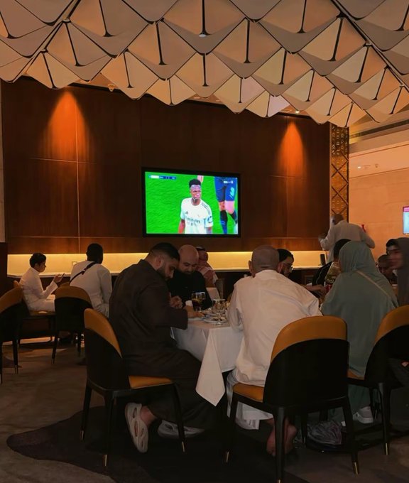 A group of people dine at a restaurant table beneath a geometric ceiling installation, watching a football match featuring a player in a white jersey on a wall-mounted TV.
