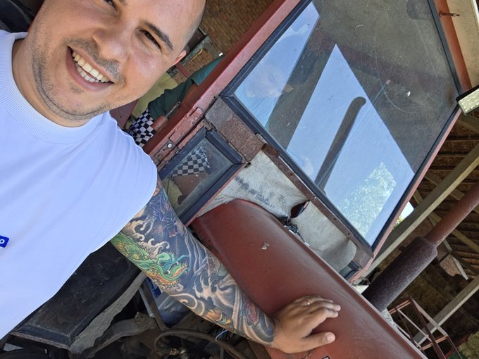 A smiling man with a colorful dragon tattoo sleeve, wearing a white t-shirt, rests his hand on a large red vessel in what appears to be a workshop.
