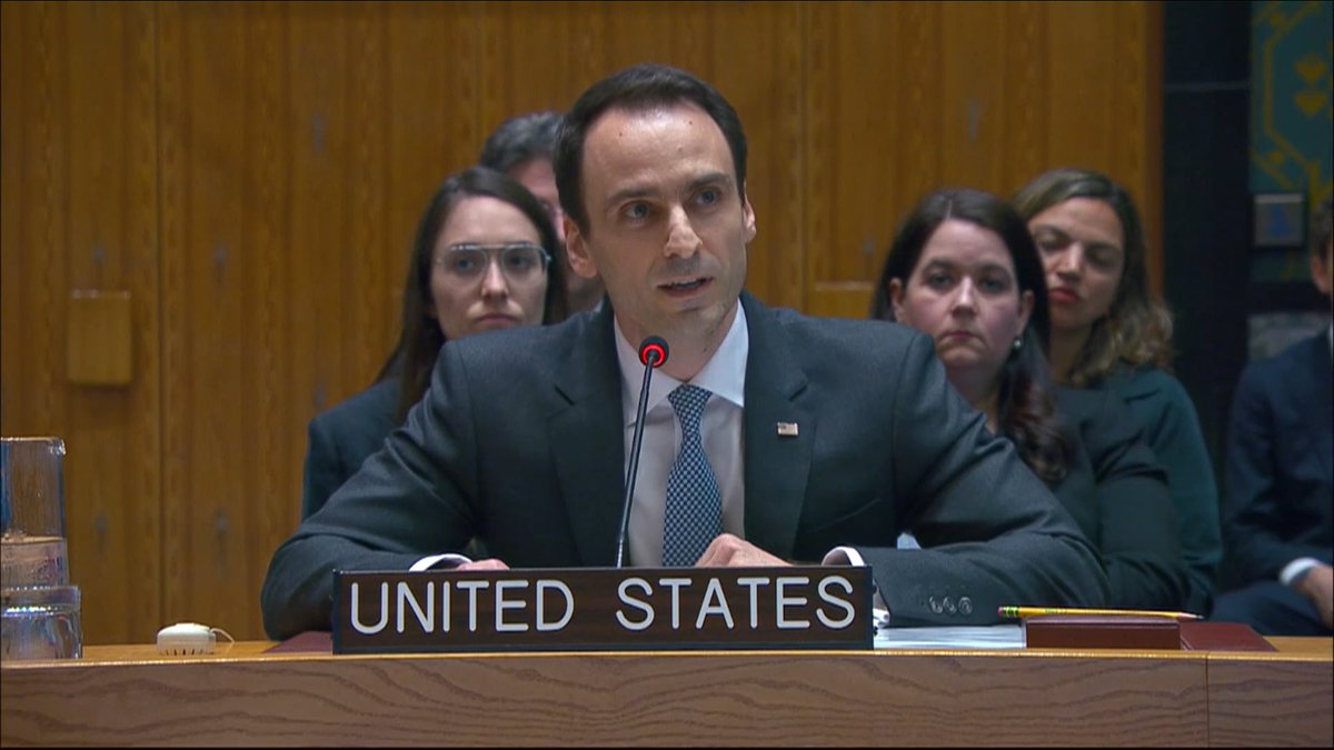 A man in a suit speaks at a microphone behind a placard reading 'UNITED STATES' in what appears to be a UN chamber, with people seated behind him.