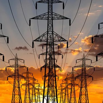 Silhouetted electrical transmission towers stretch across a vivid sunset sky, visually symbolizing the U.S. power grid facing record-high electricity costs nearing 20¢/kWh as rising demand and infrastructure investments drive consumer prices upward.